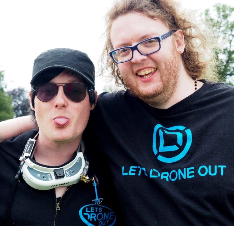AndyRC (Left) and Jack (Right) face the camera with an arm around each others shoulder. AndyRC wears FPV fatshark goggles round his neck and sticks his tongue out. Jack has a toothy smile. Both wear LDO merch; Andy in a hoodie and Jack in a t-shirt. The trees of Weston Park Model Show are visible in the background.